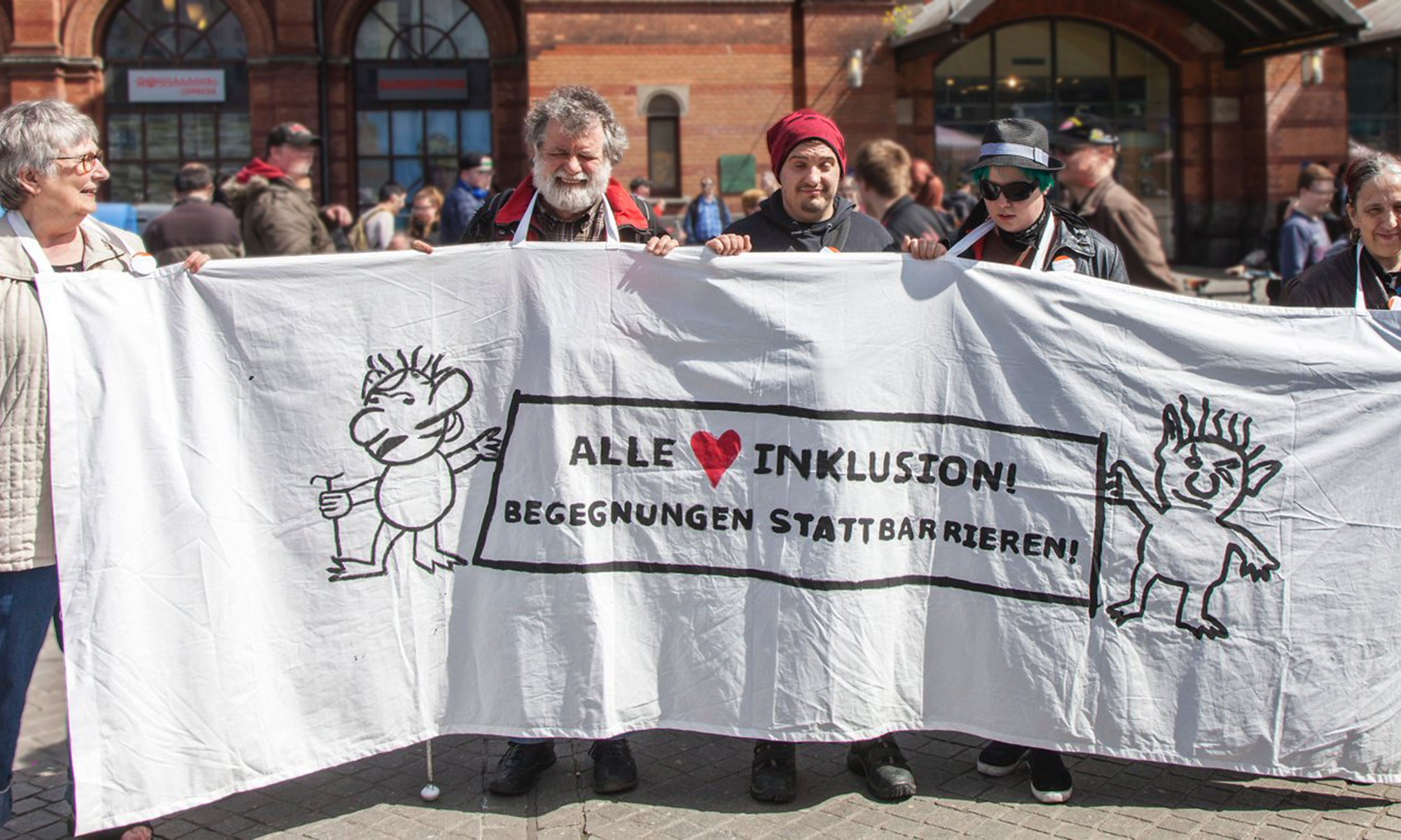 Blinde Menschen demonstrieren vor dem Hauptbahnhof mit einem Transparent auf dem steht: "Alle lieben Inklusion! Begegnungen statt Barrieren"